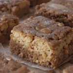 Square vanilla brownies with a light and dark layered appearance, topped with a sugary crust and scattered nuts, arranged on a wooden surface with parchment paper.